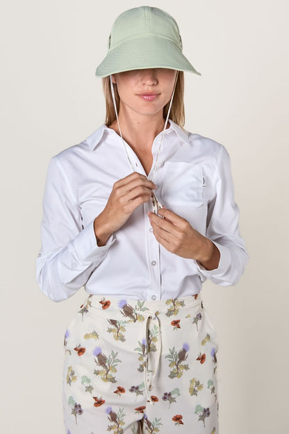 Femme portant une chemise blanche anti-UV et un chapeau vert, prête pour une protection solaire élégante.